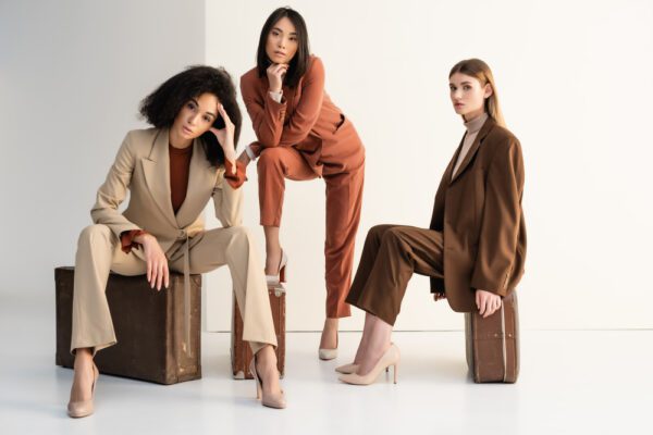 full length of stylish multicultural women in suits sitting on suitcases on white